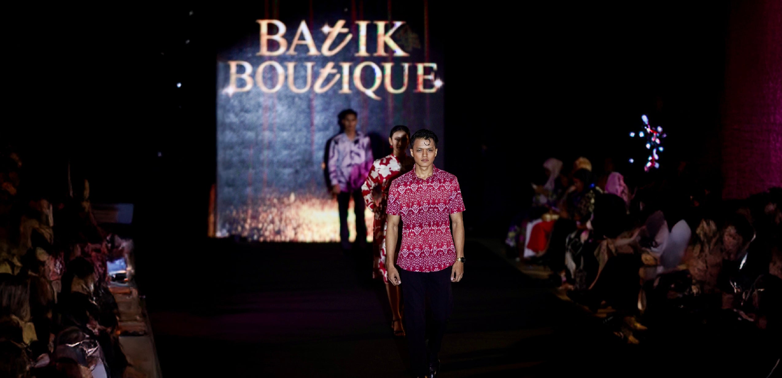 Model wearing the red Anyam batik shirt walks down the runway at the Batik Lestari Fashion Show, along with other models in the background showcasing Batik Boutique's Holiday Collection, with the Batik Boutique logo illuminated behind them.