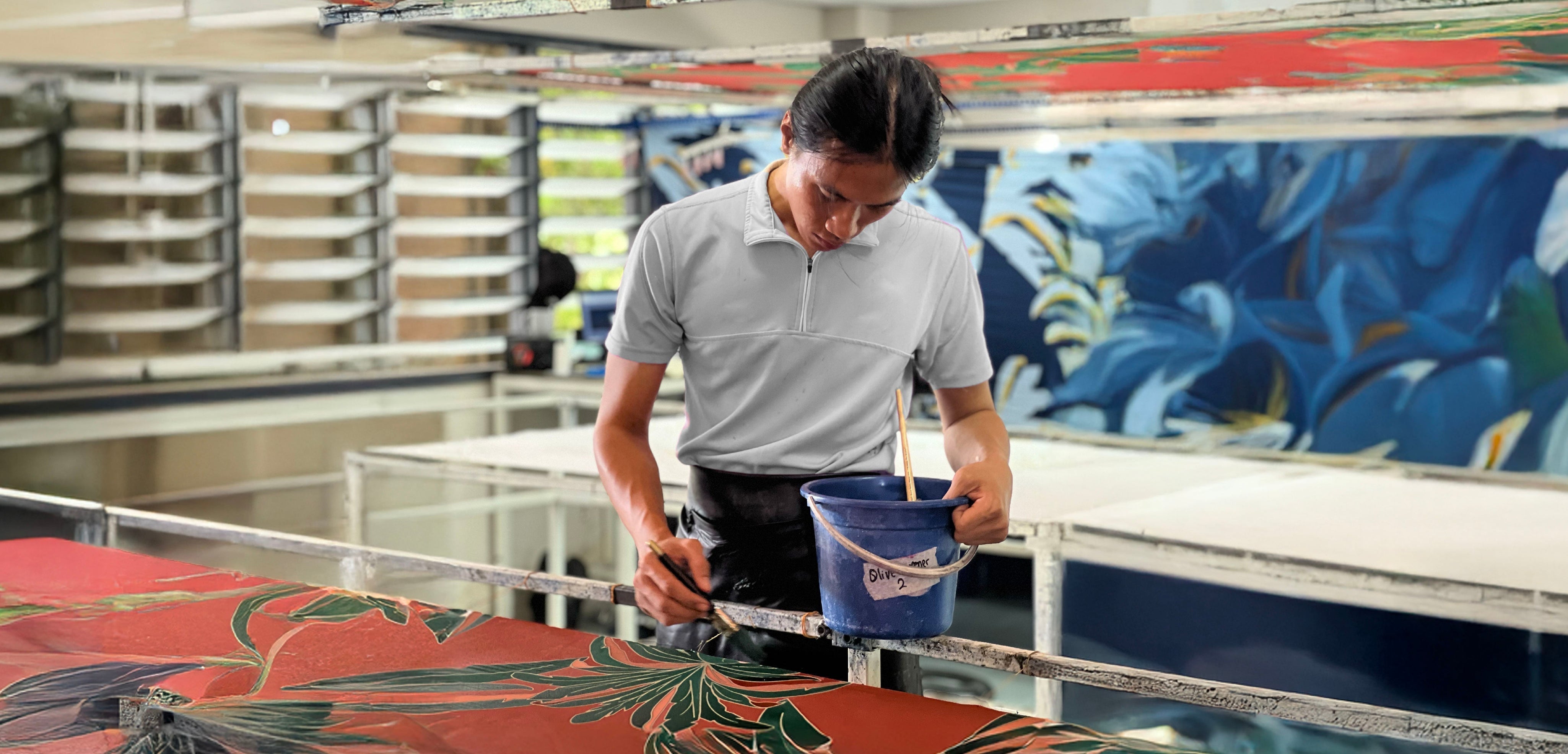 A Malaysian artisan hand-painting batik fabric at Batik Boutique’s production house. Surrounded by vibrant batik textiles, the artisan carefully applies color, showcasing the precision and artistry of traditional batik-making.