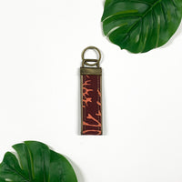 a lifestyle photo of a keyfob made of batik in the pattern maroon coral against a neutral background surrounded by two tropical plants