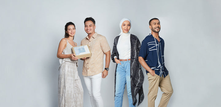 Two couples standing in front of white background with a gift box. One couple is wearing pastel batik apparel and the other is wearing bold and modern batik apparel.