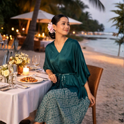 Woman in a green dress sitting at a beachside dinner table with palm trees and water in the background.