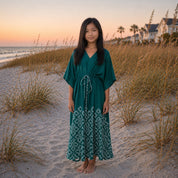 Young girl in a green dress standing on a sandy beach with grasses and houses in the background.
