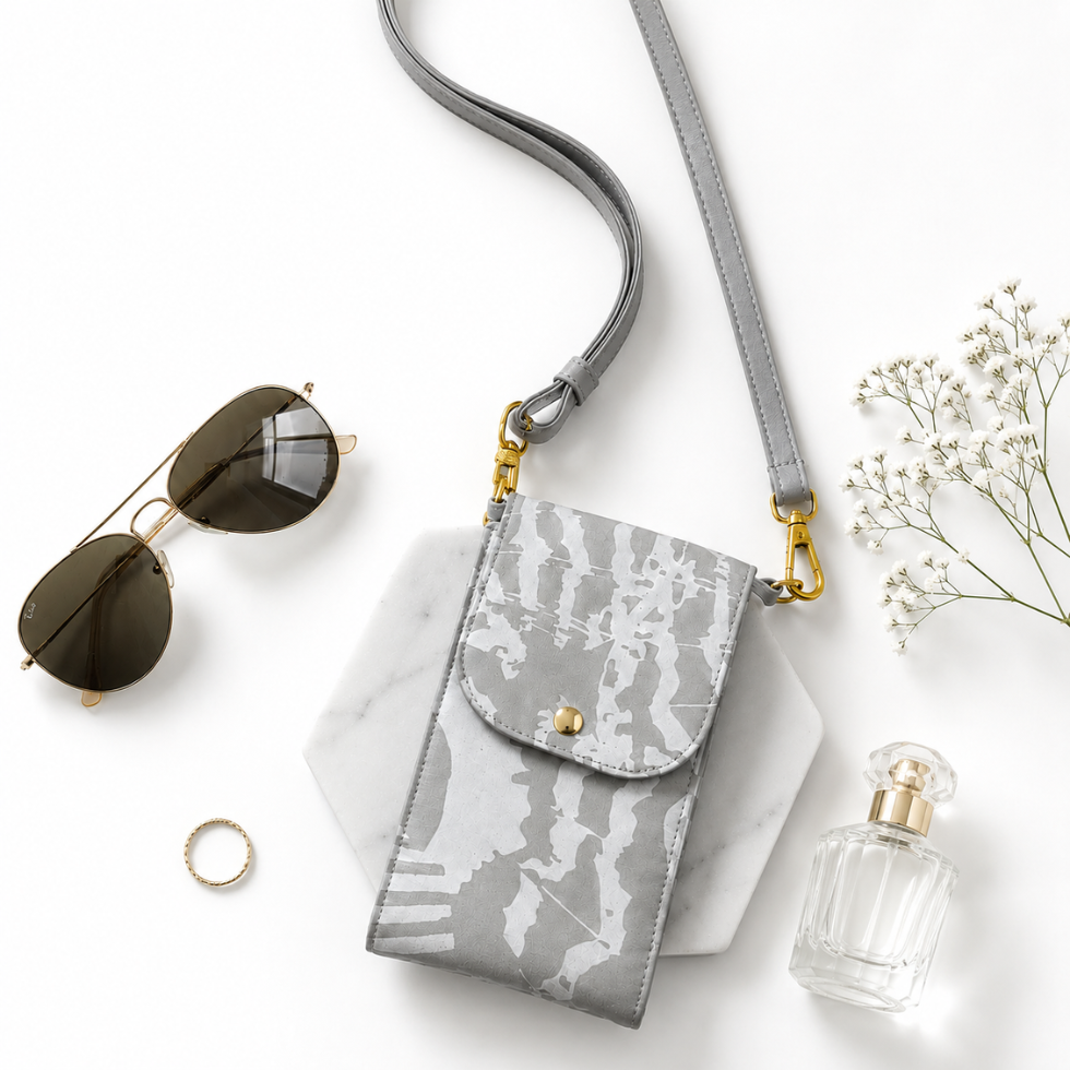 A gray crossbody phone bag with a tie-dye patterned flap and gold hardware, styled in a flat lay with sunglasses, a ring, a perfume bottle, and baby's breath on a white background.