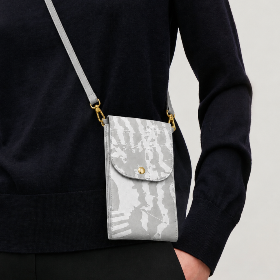 A person wearing a black sweater carries a small gray crossbody phone pouch with a gray abstract pattern, gold hardware, and a gold snap button.