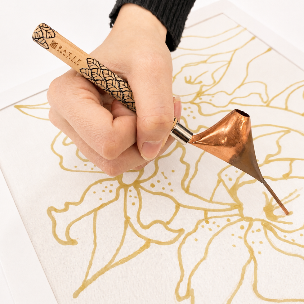 A close-up of a hand holding a Nafis brand copper calligraphy tool over white paper, drawing a gold floral pattern.