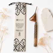 A handmade copper and wood hand tool with a black patterned handle, displayed next to its beige drawstring pouch with black batik leaf patterns. The set is styled on a white background with dried flowers and a stone.