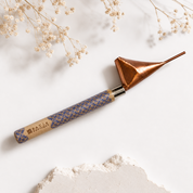 A handcrafted metal and wood tool with a copper-colored conical head, a silver collar, and a patterned blue and gold handle, styled on a white background with dried flowers and a stone slice.