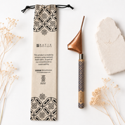 A handcrafted batik clay spoon with a copper-colored metal bowl and a patterned wooden handle, displayed next to its branded kraft paper pouch on a white background with dried flowers and a stone.