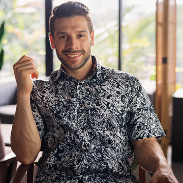 in a lifestyle photo, a male model is posing while sitting on a chair posing in an authentic batik shirt in the pattern black floret