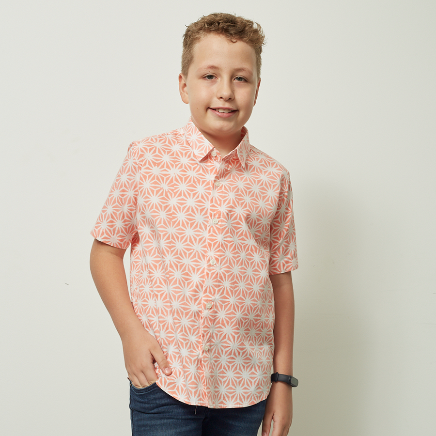 A young boy confidently posing against a neutral background, proudly showcasing the Peach Firework batik attire he's wearing