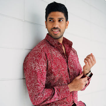 A man is leaning on white wall wearing red handmade color batik long sleeve in cotton material. 
