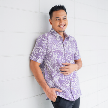 a man leaning against a white wall while wearing a malaysian batik shirt in the pattern lavender bintik