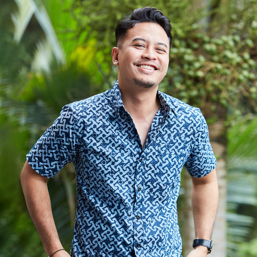 A man wearing blue batik shirt in the pattern midnight arabesque standing in front of greenery