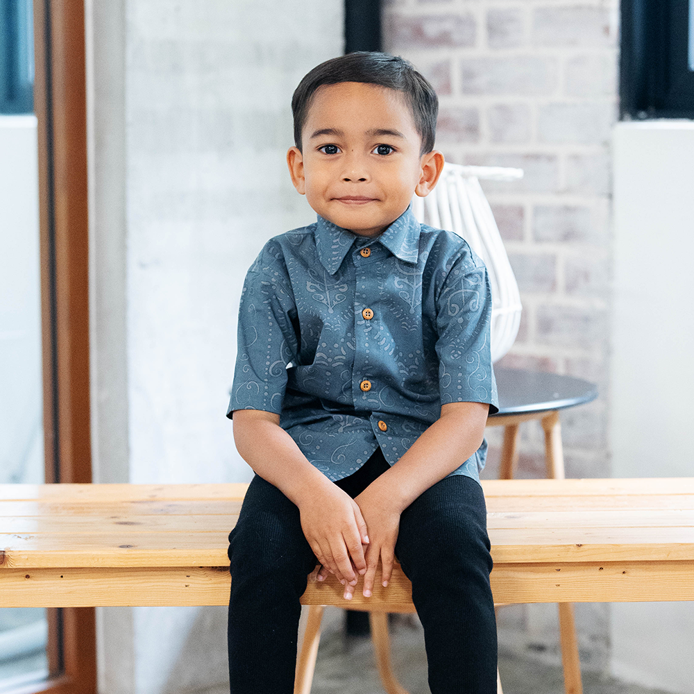 A boy sitting on a bench wearing a short-sleeved blue batik shirt with collar and buttons, paired with black pants.
