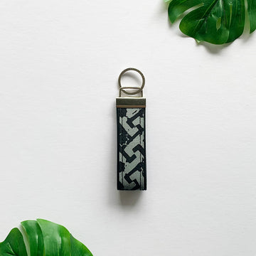 a lifestyle picture of a key fob made of batik in the patter grey arabesque against a neutral background and with tropical leaves as decorations