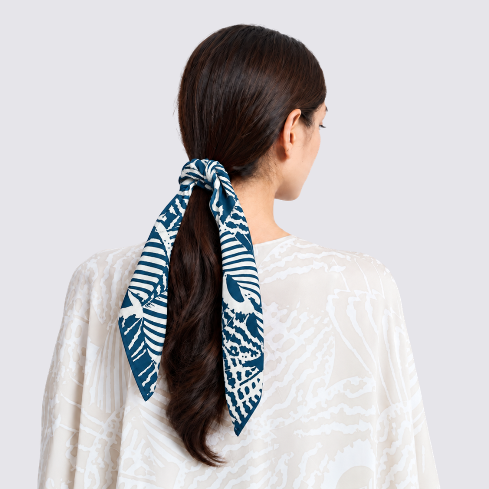 A woman shown from the back wearing a navy blue and white patterned silk scarf tied around her dark brown ponytail, paired with a white patterned top against a light gray background.