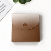 A square kraft paper envelope-style box with a flap closure and a small circular cutout detail, photographed on a clean white surface with a book corner and green leaves as styled props.