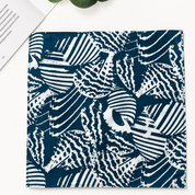 A square fabric item, likely a napkin or pocket square, featuring a navy blue and white abstract block-print pattern. It is presented flat on a white surface with a book corner and a green leaf visible in the top-left.