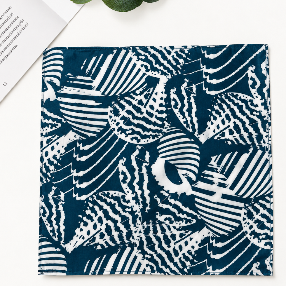 A square fabric item, likely a napkin or pocket square, featuring a navy blue and white abstract block-print pattern. It is presented flat on a white surface with a book corner and a green leaf visible in the top-left.