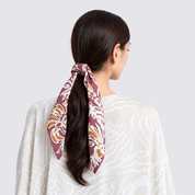 A woman shown from the back wearing a white patterned top and a dark brown hair tie with a burgundy, gold, and white floral scarf, set against a light gray studio background.