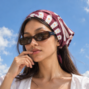 A woman outdoors under a blue sky wearing a burgundy and white floral headscarf and dark sunglasses.