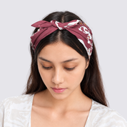 A woman wearing a burgundy and white patterned silk headband with a bow, looking down against a light gray background.