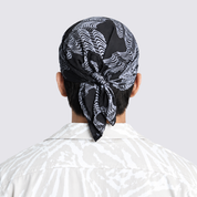 A man viewed from behind wearing a black and white patterned silk headscarf tied in a knot at the back of his head, paired with a matching abstract-patterned shirt against a light gray background.