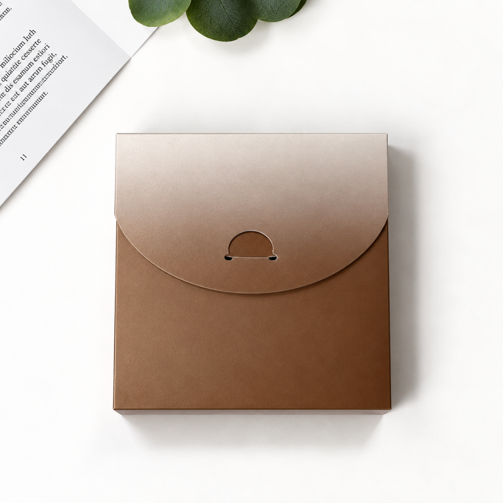 A square kraft paper envelope-style box with a flap closure and a small circular cutout detail, photographed on a clean white surface with a book corner and green leaves as styled props.