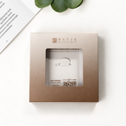 A square metallic taupe packaging box with a clear window displaying a small white jewelry stand, branded 'BATIK BOUTIQUE' in gold lettering. The box is photographed from above on a clean white surface with a green leaf and a corner of a printed page as styled props.