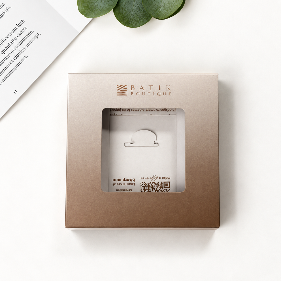 A square metallic taupe packaging box with a clear window displaying a small white jewelry stand, branded 'BATIK BOUTIQUE' in gold lettering. The box is photographed from above on a clean white surface with a green leaf and a corner of a printed page as styled props.