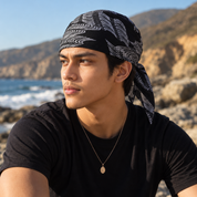 A man wearing a black and white patterned headwrap bandana sits outdoors on a rocky beach with the ocean and hills in the background.