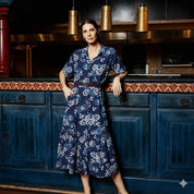 Woman in a blue floral dress standing in a kitchen with dark wooden cabinets and pendant lights.