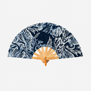 A folding hand fan with a navy blue and white batik pattern on the fabric, featuring a light wood bamboo frame and a small metal brand plaque that reads 'M. LATTIE'.