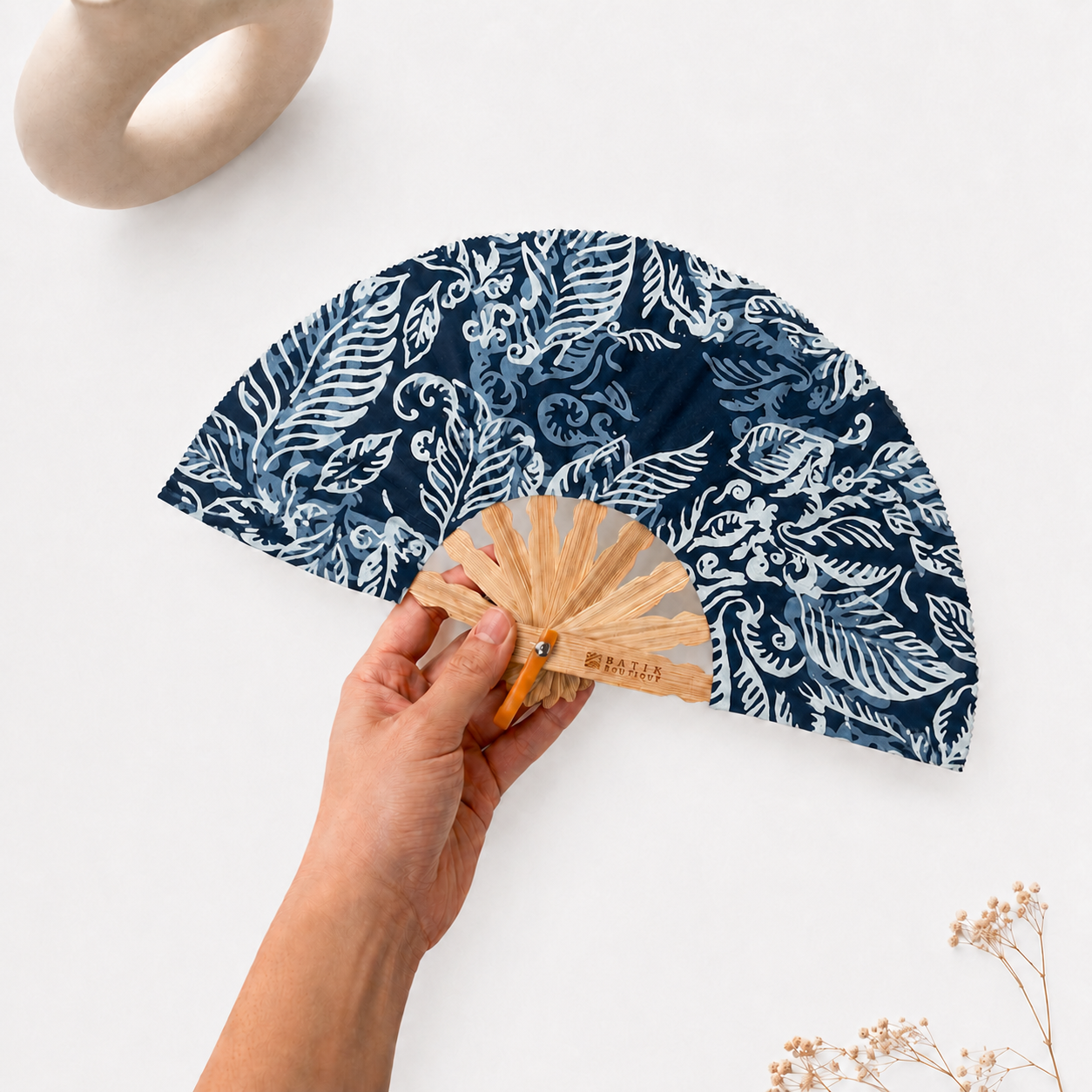 A hand holds a folding hand fan with a navy blue and white botanical pattern. The fan has a light natural wood handle and a small orange accent near the hinge.