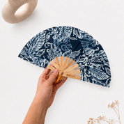 A hand holds a folding hand fan with a navy blue and white botanical pattern. The fan has a light natural wood handle and a small orange accent near the hinge.