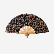 A folding hand fan with a black and beige abstract web-like patterned fabric, a light natural wood handle, and a small metal brand plaque reading 'SOUVENIR'.