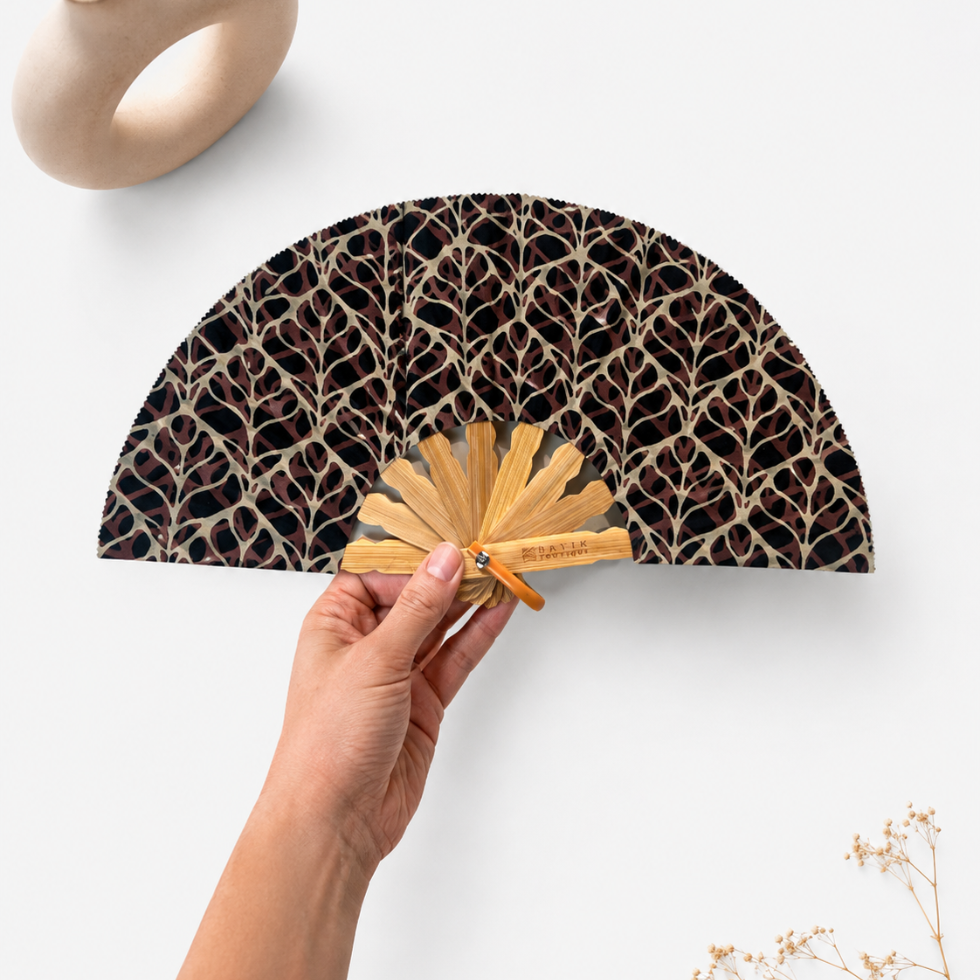 A hand holds a folding hand fan with a black background and a beige leaf pattern. The fan has a natural bamboo handle and a small orange wrist strap.