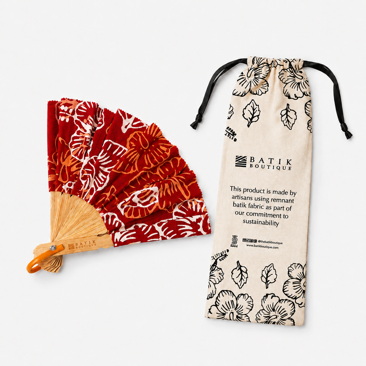 A red and orange batik paper fan with a wooden handle lies next to a beige drawstring pouch printed with black floral motifs and the text 'BATIK BOUTIQUE'.