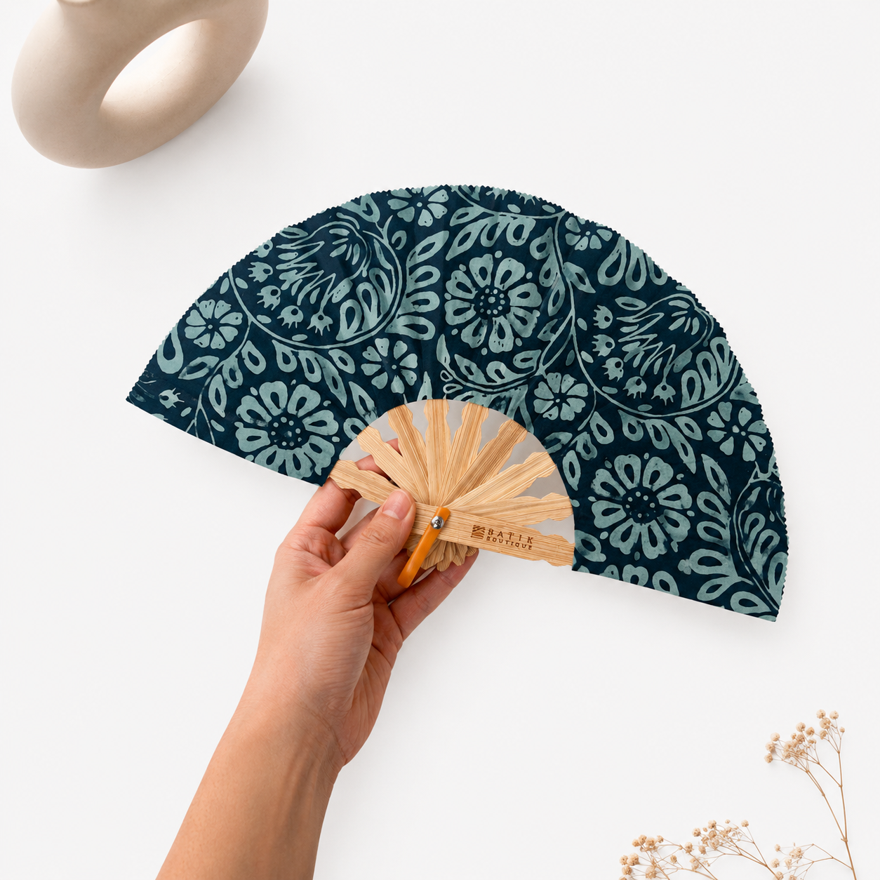 A hand holds a folding hand fan with a dark teal and light blue floral patterned fabric. The fan has a natural bamboo handle with the brand name 'MAJIA' printed on it.