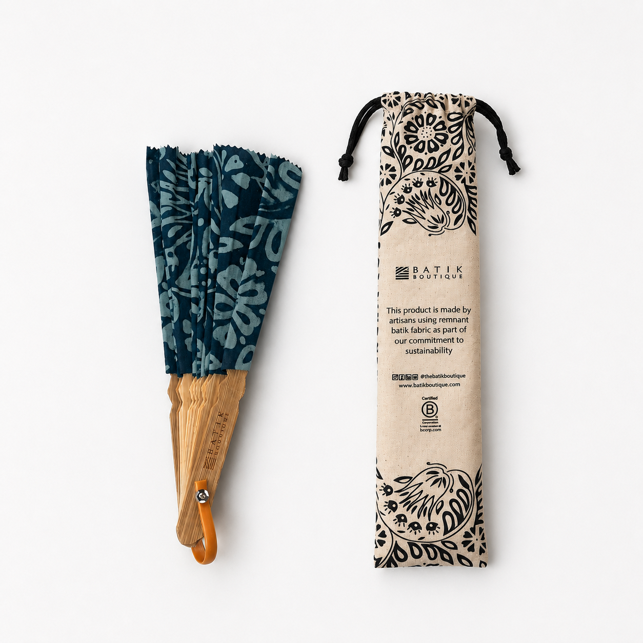 A folded batik-style hand fan with a dark teal floral pattern and light wood handle, shown next to its beige drawstring pouch printed with black floral motifs and Patik Boutiques branding, on a clean white background.