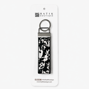 A Batik Boutique keychain with a black and white floral ikat pattern on a fabric strap, attached to a silver metal ring and hanger, displayed on a white backing card.