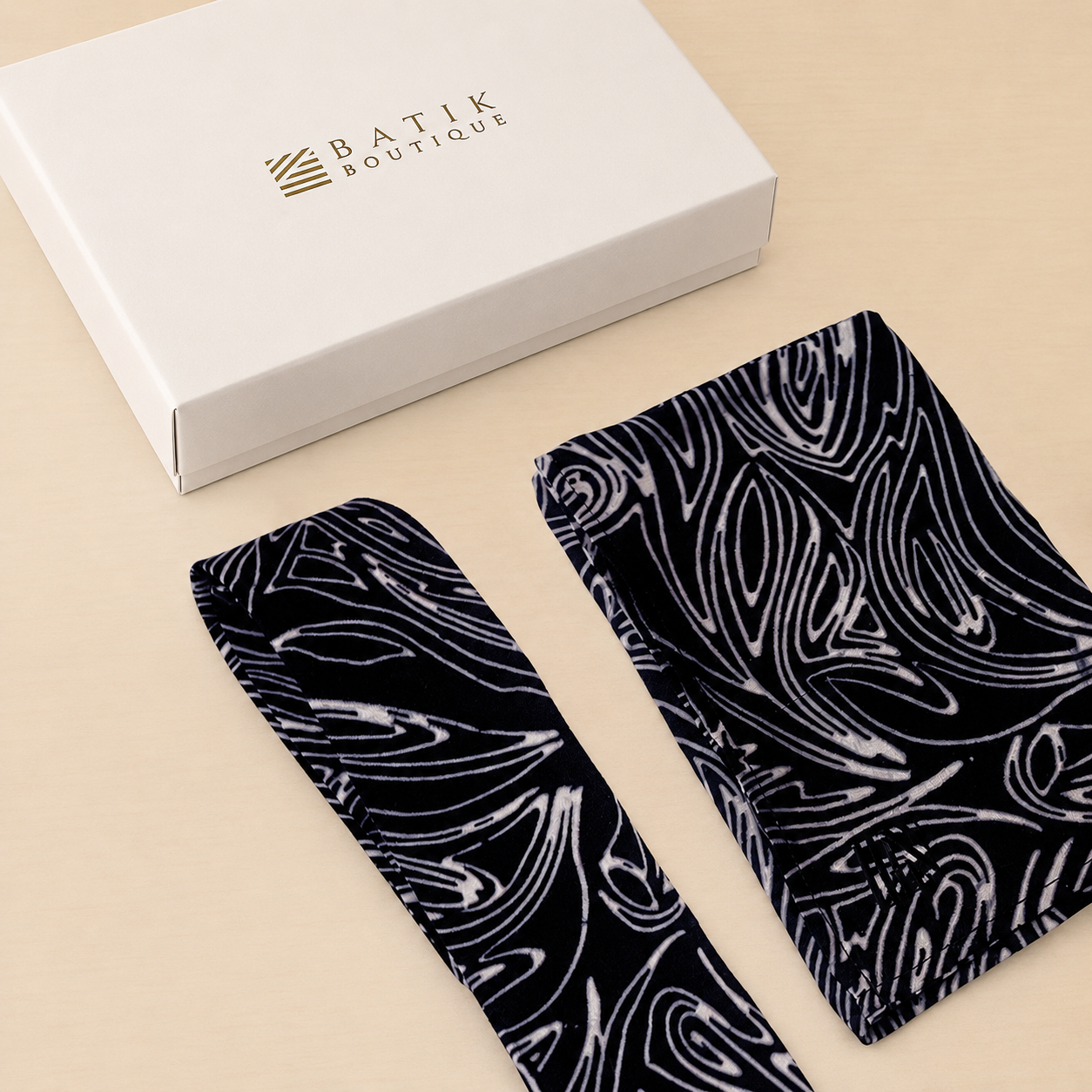 Two black batik fabric accessories with white abstract wave patterns are displayed next to a white 'BATIK BOUTIQUE' box on a light beige background.