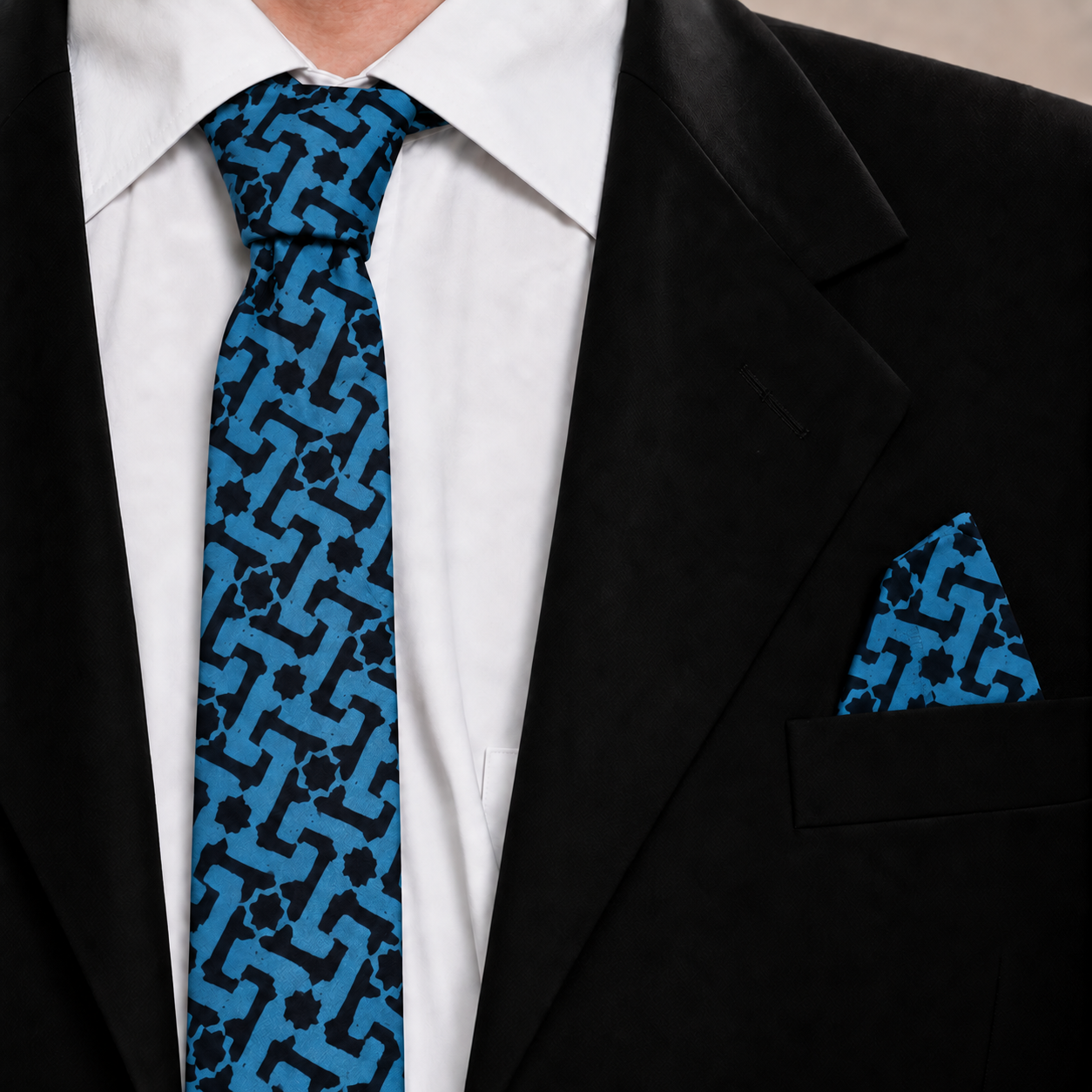 A close-up of a men's necktie and matching pocket square set in a black suit jacket and white dress shirt. The tie features a geometric interlocking pattern in bright blue and black.