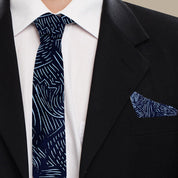 A close-up of a men's necktie and matching pocket square set, featuring a navy blue and white abstract wavy line pattern, styled on a white dress shirt and black suit jacket.