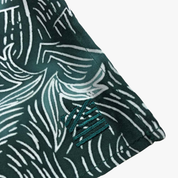 Close-up of a dark green and white patterned fabric item, showing a folded corner with a small embroidered logo patch on a light gray background.