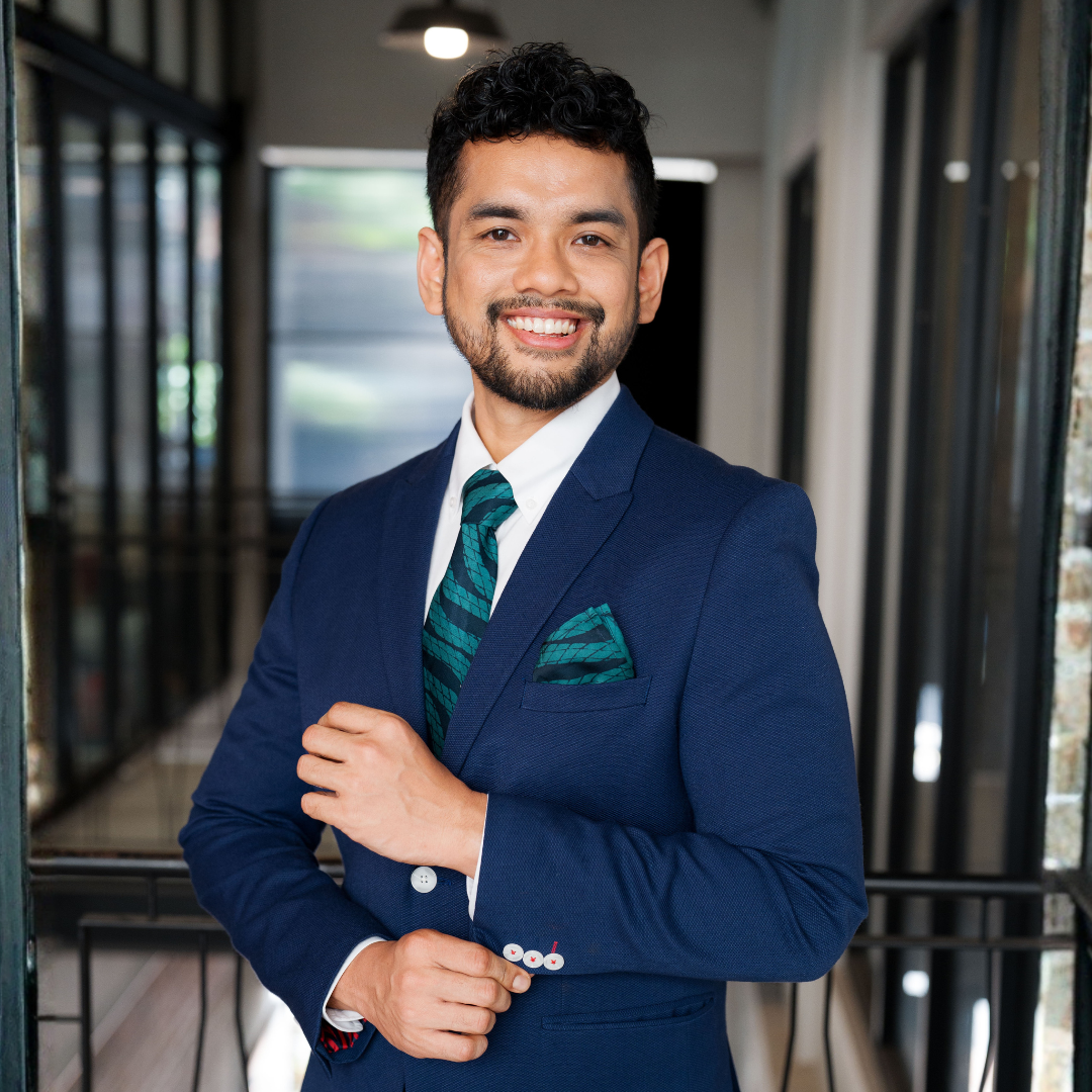 Man in a blue suit with a green tie and pocket square standing in an indoor setting.