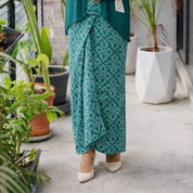Person wearing a green outfit with a patterned skirt standing in an outdoor setting with plants.