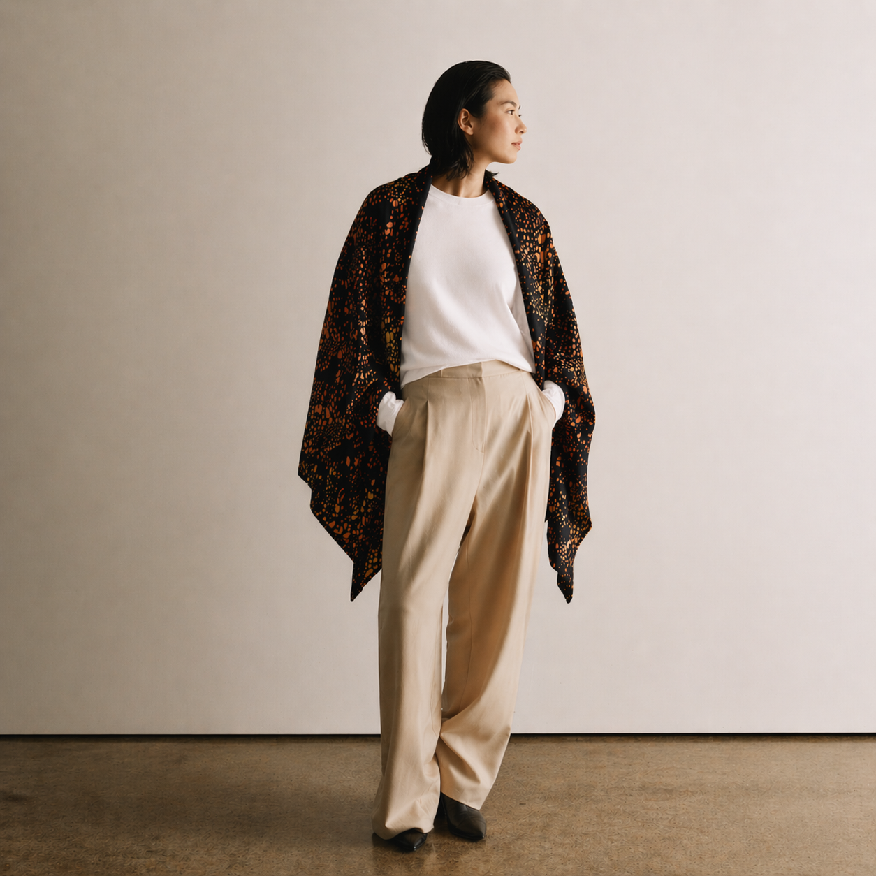 Person wearing a patterned shawl, white shirt, beige pants, and black shoes against a plain background
