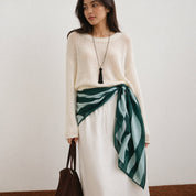 Woman wearing a white sweater and white skirt with a green and blue scarf, holding a brown bag.