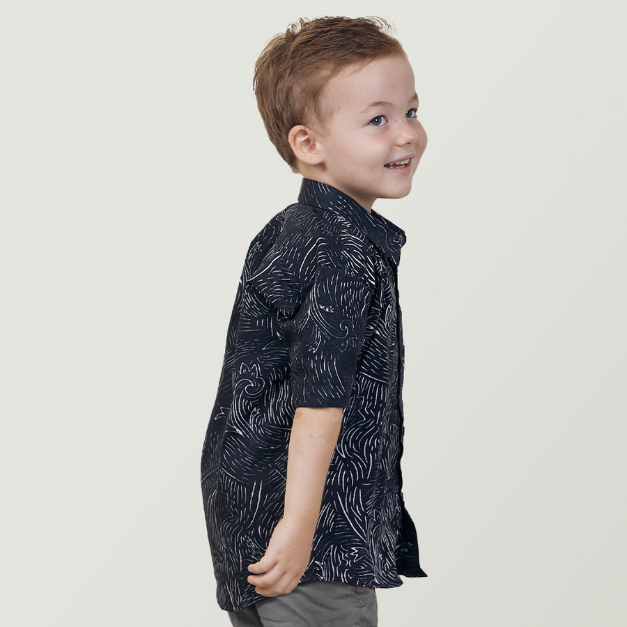 a boy looking over his shoulder while standing in front of a white wall in a black driftwood batik shirt